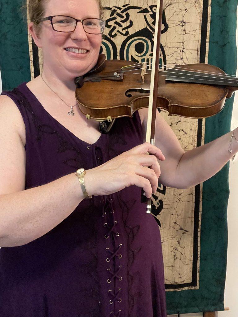Woman holds violin and shows her fiddle bow hold with her hand holding the end of the bow near the frog in front of a Celtic banner.