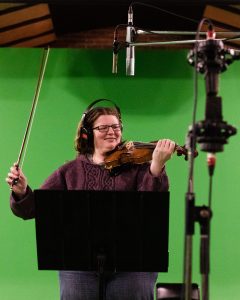 Katherine Moller pictured in a studio, against a green screen with fiddle in hand and a microphone in front of her. She has a relaxed and joyful posture.