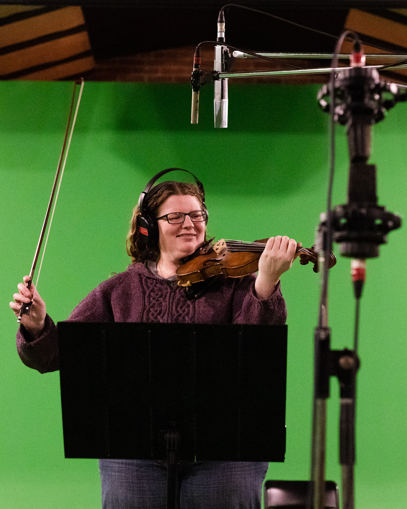 Katherine Moller pictured in a studio, against a green screen with fiddle in hand and a microphone in front of her. She has a relaxed and joyful posture.